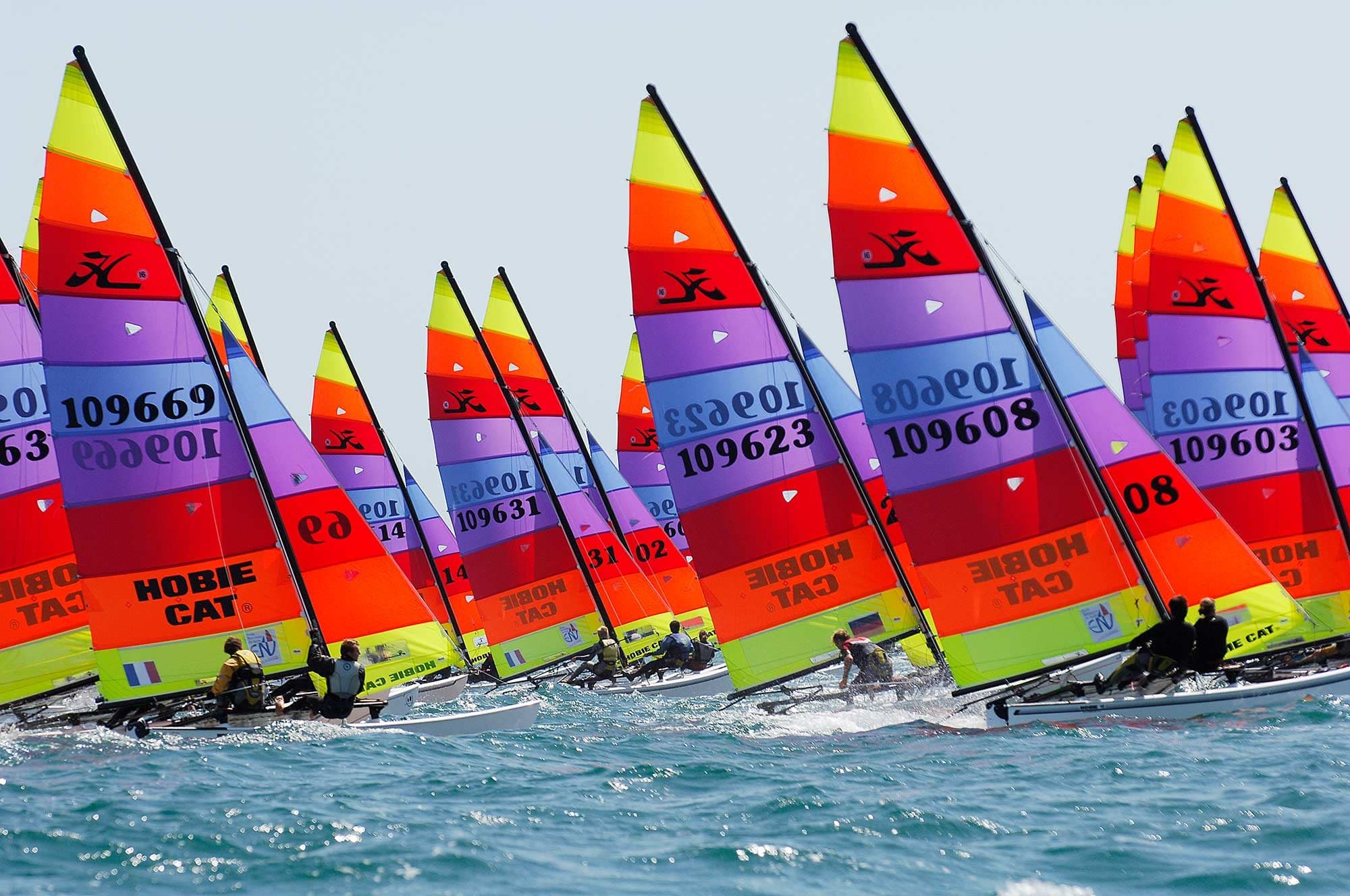 A fleet of Hobie 16 catamarans with rainbow-striped sails racing side by side